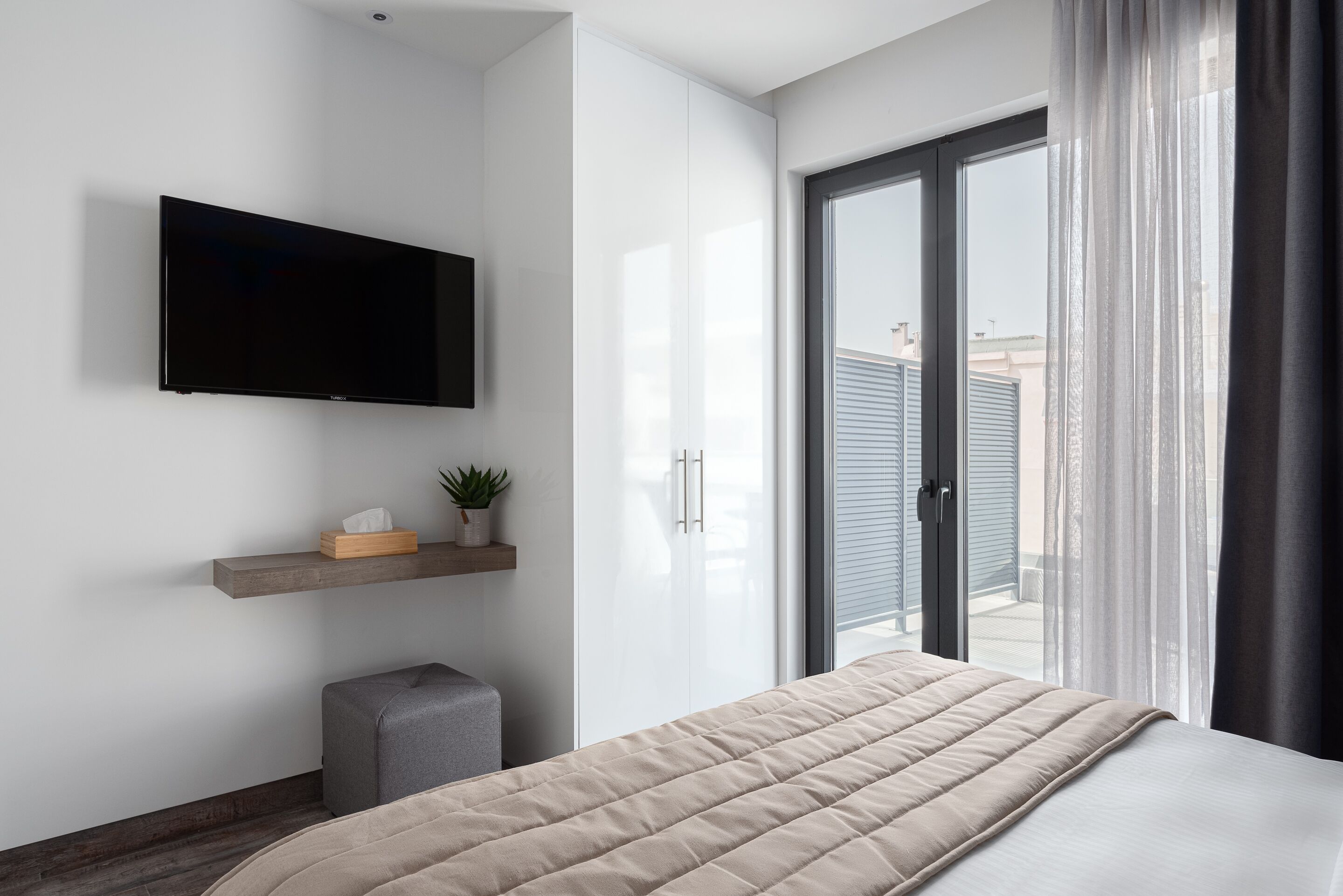 A modern bedroom with a bed, a mounted TV, a small plant, and a sliding door to a balcony.