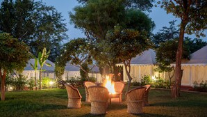 A fire pit surrounded by wicker chairs in a grassy area with trees and tents in the background. - Ranthambore Tiger Resort by Park Tree (Sawai Madhopur)