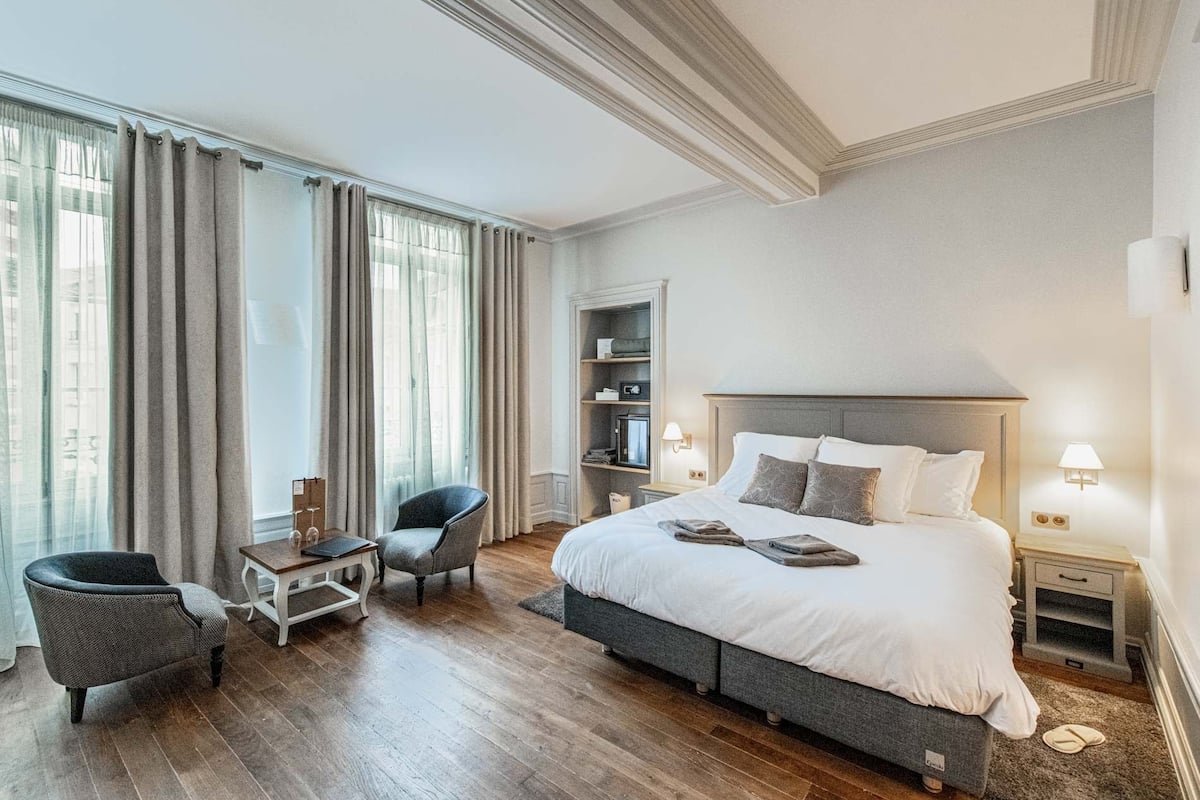Une chambre d’hôtel avec un grand lit, deux fauteuils, une petite table, une bibliothèque et une fenêtre avec des rideaux.