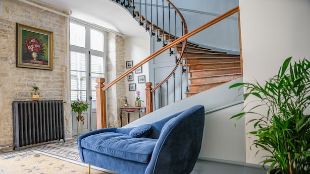 A modern living room with a blue sofa, a staircase with wooden railings, a radiator, and a potted plant. - Maison Qualisterra - Chambre d'Hôtes (Bar-sur-Aube)