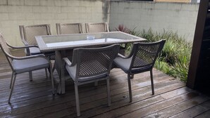 Un patio con una mesa de vidrio y sillas de mimbre, rodeado por una terraza de madera y un muro de hormigón.