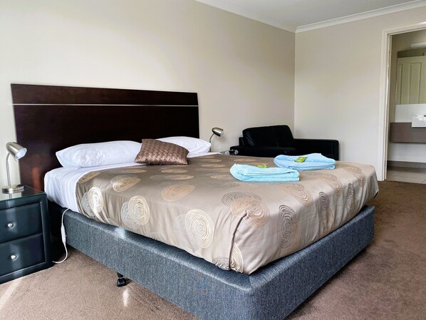 A neatly made bed with a patterned bedspread, a bedside lamp, and a black chair. - Dalvue Motel (Terang)