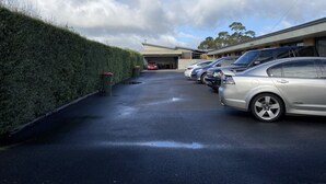 Un estacionamiento con varios coches aparcados en fila, un seto a la izquierda y un edificio al fondo.