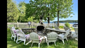An outdoor seating area with stone fire pit and white chairs, surrounded by a white picket fence, overlooking a lake.