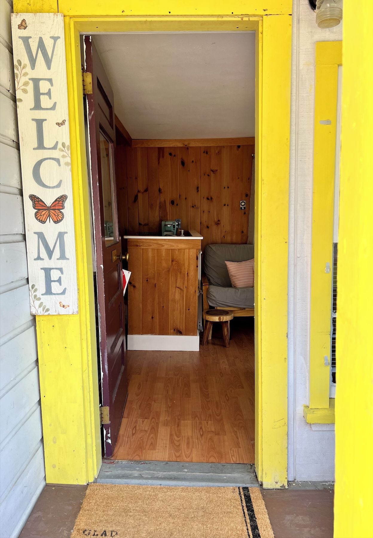 A wooden interior with a 'WELCOME' sign, a small stool, and a couch.