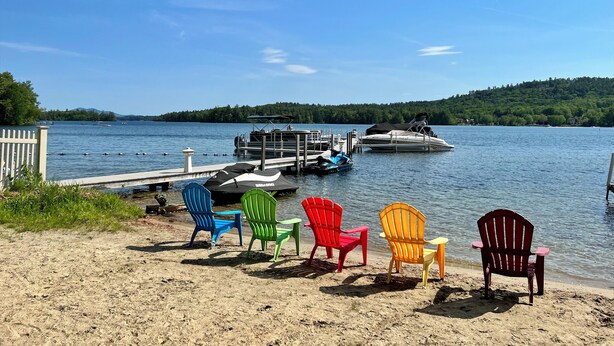 A lakeside view with a dock, several boats, and colorful Adirondack chairs on the sandy shore.
