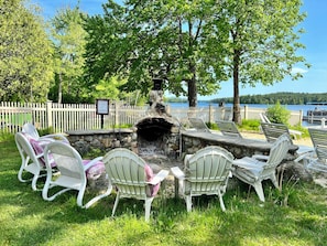 An outdoor seating area with stone fire pit and white chairs, surrounded by a white picket fence, overlooking a lake.