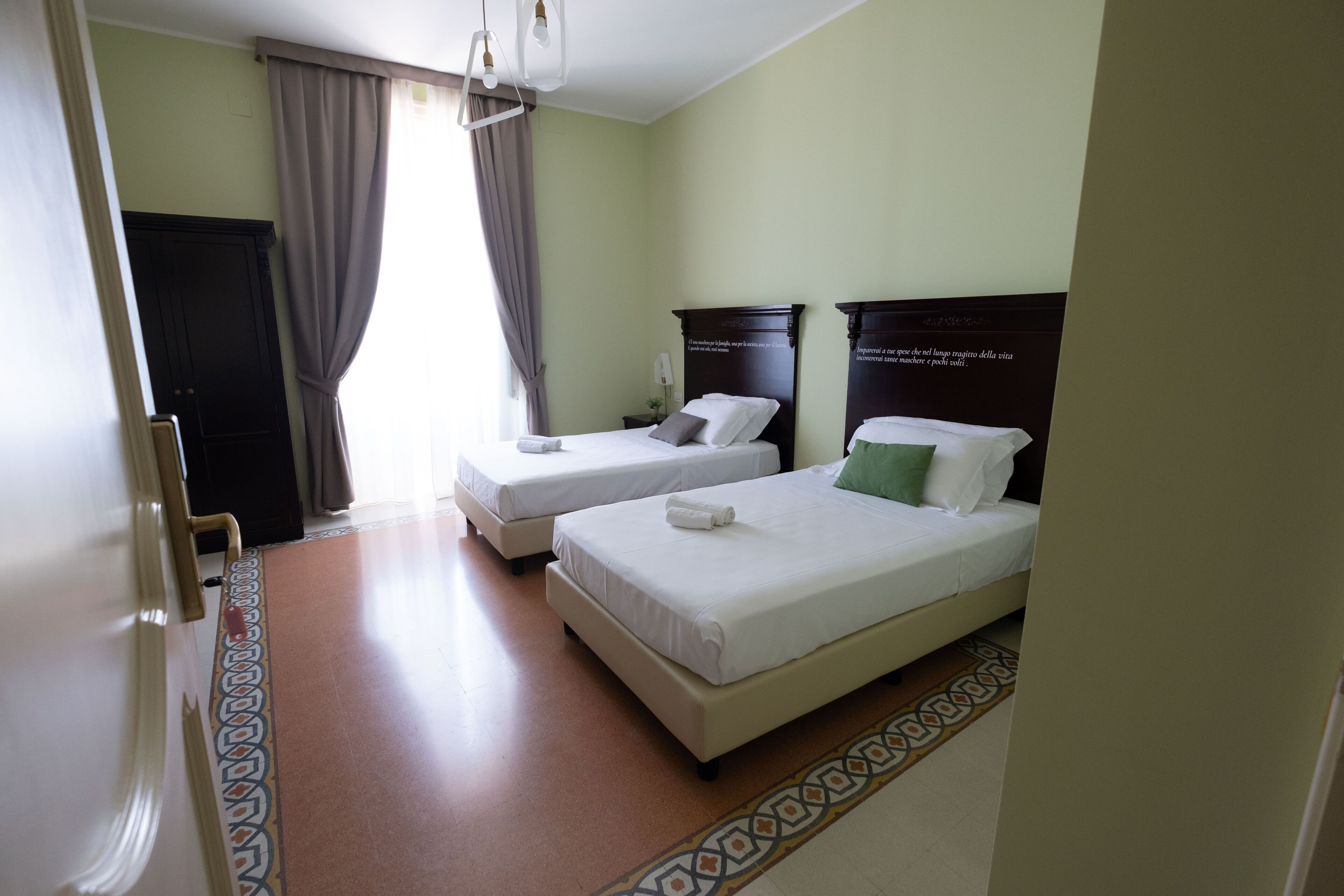 A hotel room with two single beds, a large window with curtains, a patterned floor, and a dark wooden headboard.
