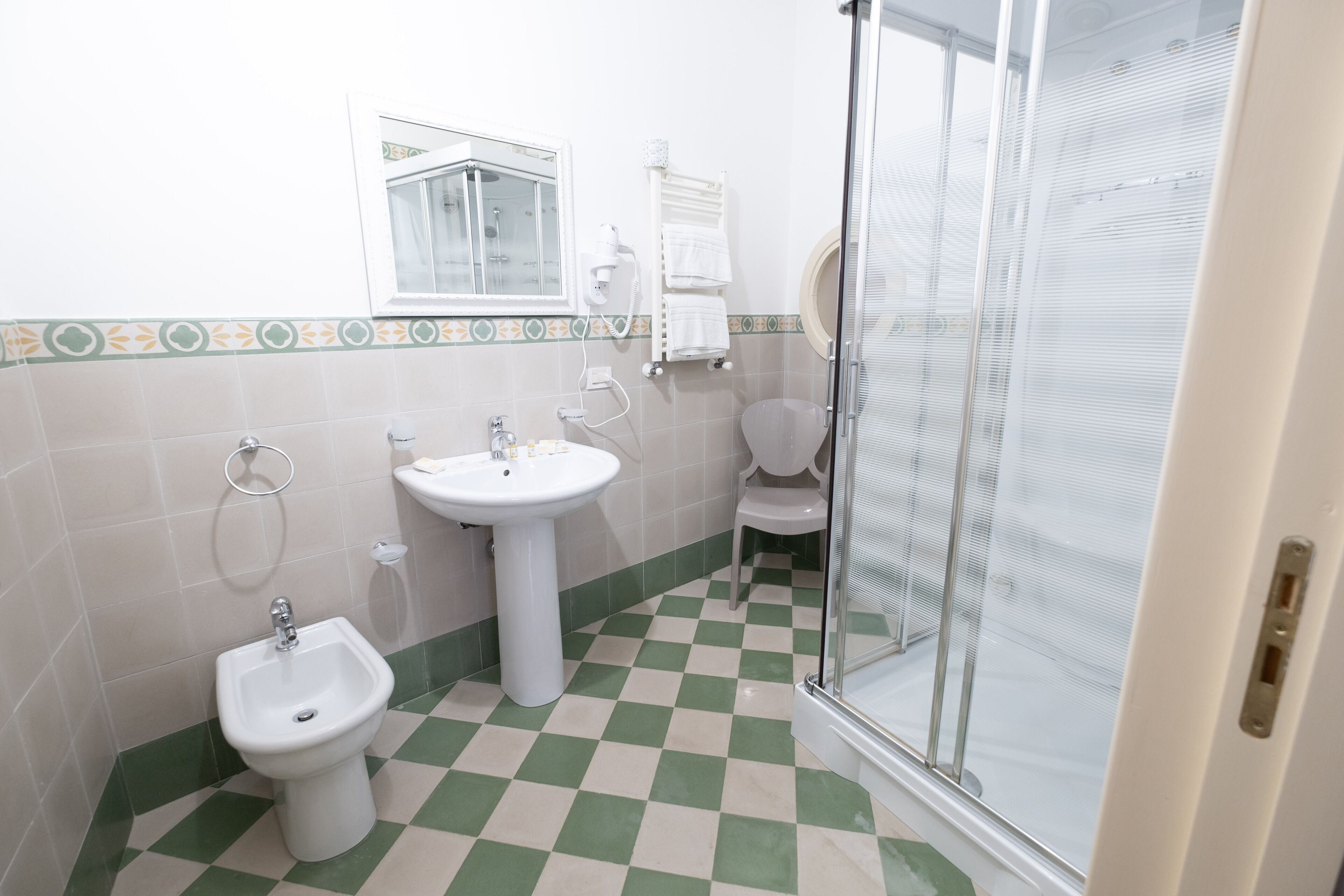 A bathroom with a bidet, sink, and toilet, featuring checkered floor tiles and a window.
