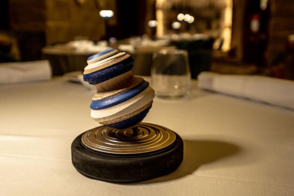 A wooden object with a spiral design on a black base, placed on a table with a glass of water in the background.