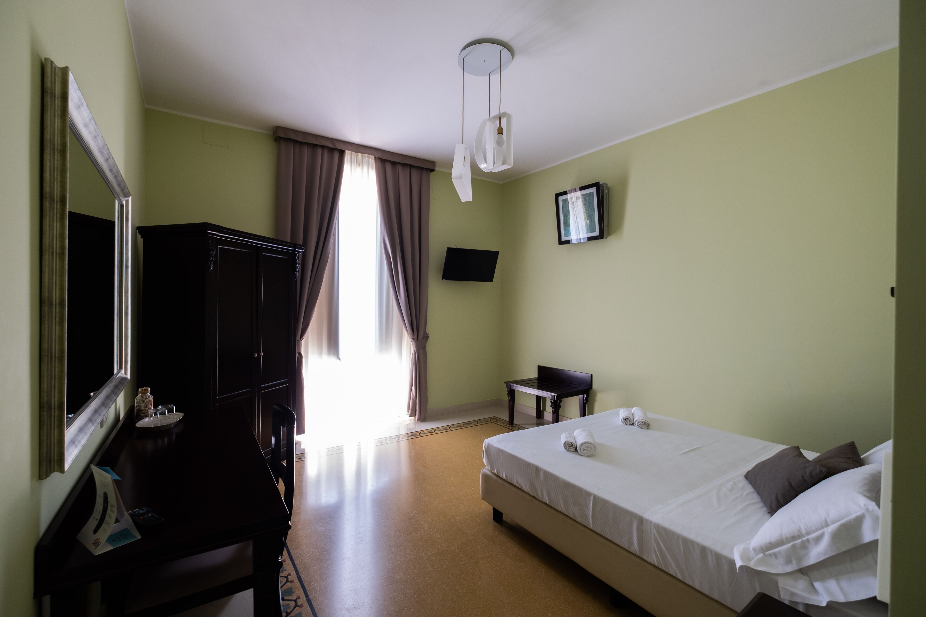 A hotel room with a bed, a desk, a chair, a dresser, a mirror, a TV, and a window with curtains.
