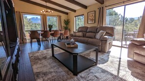 A spacious living room with a large sofa, a coffee table, and a dining area with chairs and a table. There are large windows providing a view of the outdoors. - Sedona 4BR • Red Rock Views • Deck + Game Room (Sedona)