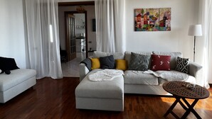 A modern living room with a grey sectional sofa, a round wooden coffee table, and a white armchair. A colorful abstract painting is on the wall, and the room has white curtains. - Private garden, tranquility and privacy (San Michele-montecchio)