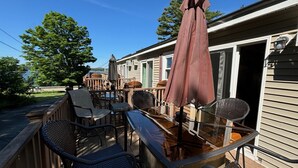 A deck with outdoor furniture, including chairs and a table, and a large umbrella. - Hi Spot Studio with shared beach on Winnipesaukee! (Laconia)