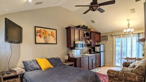A bedroom with a bed, a nightstand, a desk, a chair, a ceiling fan, a kitchen area, and a sliding glass door leading to an outdoor area. - Hi Spot Studio with shared beach on Winnipesaukee! (Laconia)