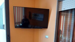 A room with a flat-screen TV mounted on the wall, a sliding glass door with curtains, and a wicker chair with a cushion. - FURNISHED TWO-ROOM APARTMENT 200 MT FROM THE LIDO OF ALGHERO (Alghero)