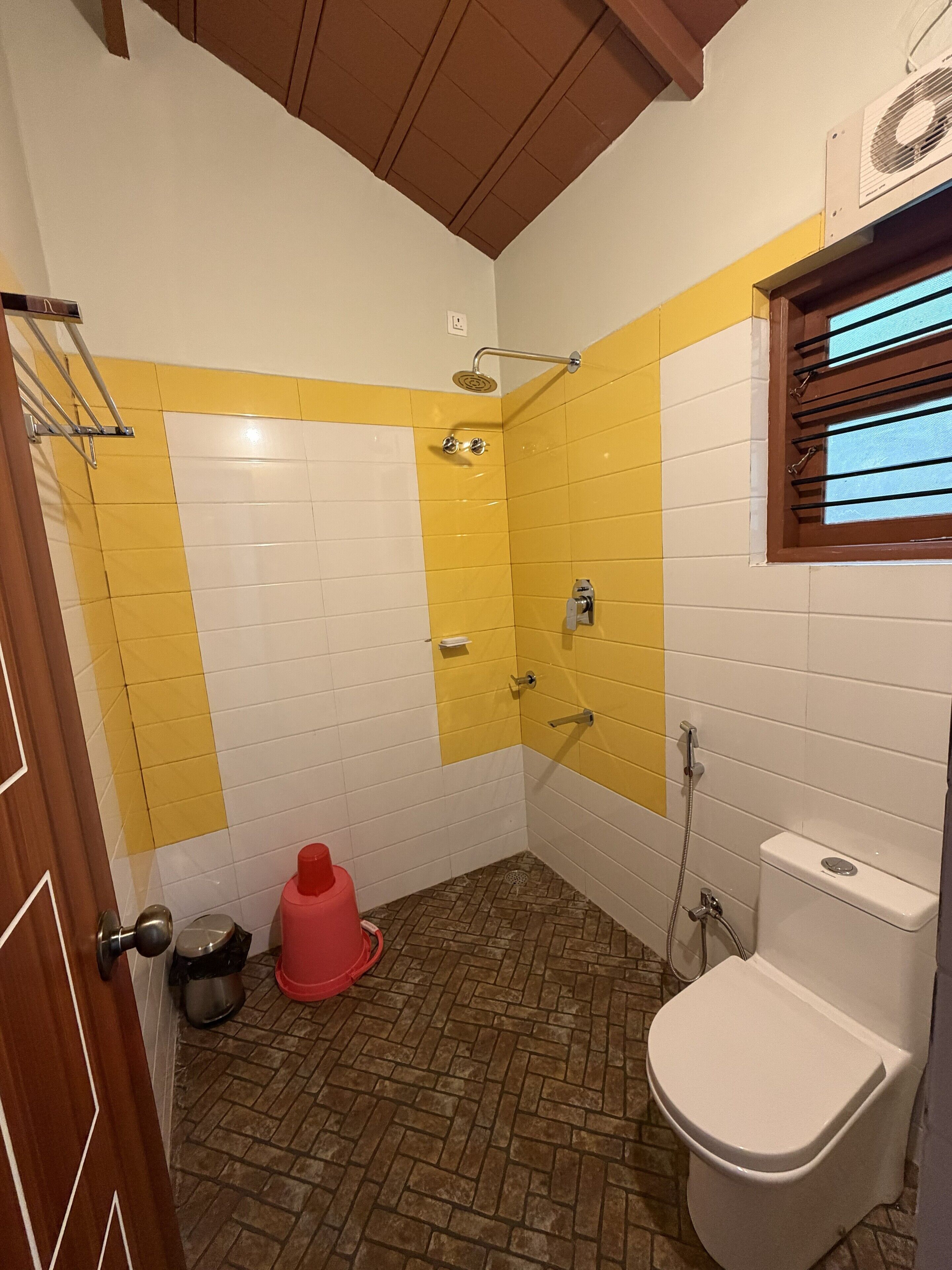 Un baño con azulejos amarillos y blancos, ducha, inodoro y ventana con persianas.