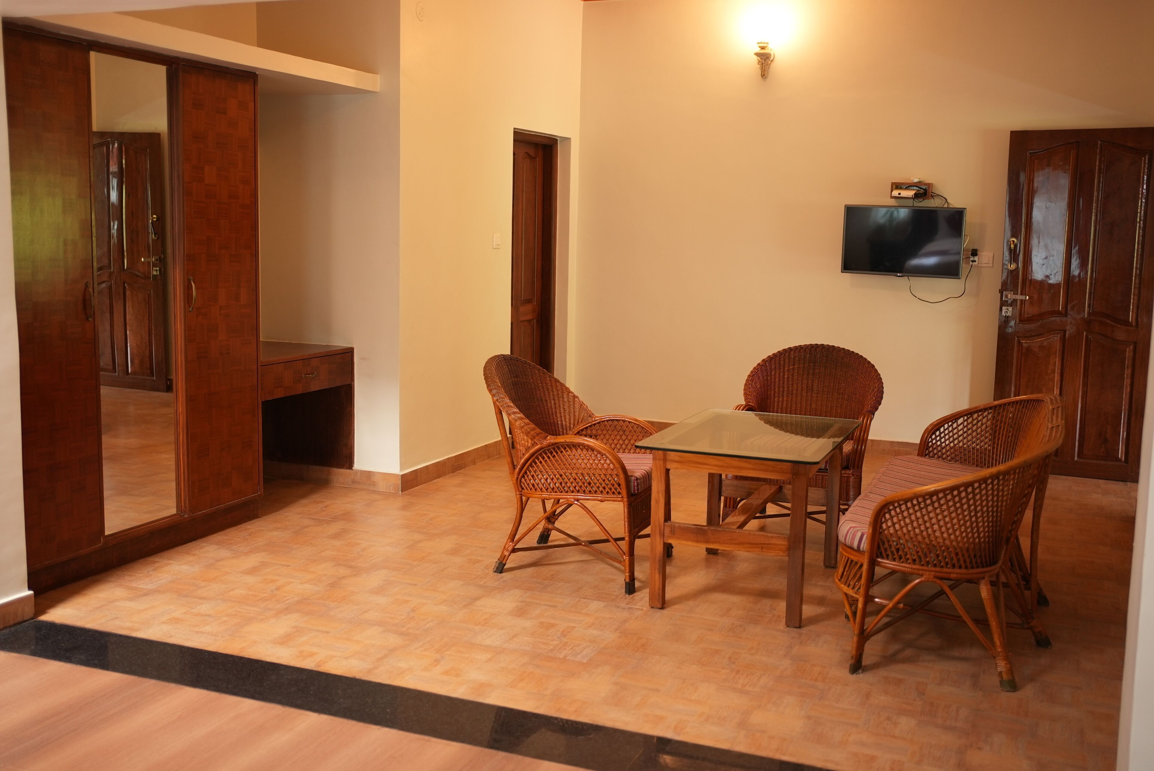 A room with a wooden floor, a glass table, wicker chairs, a television mounted on the wall, and a wooden wardrobe.