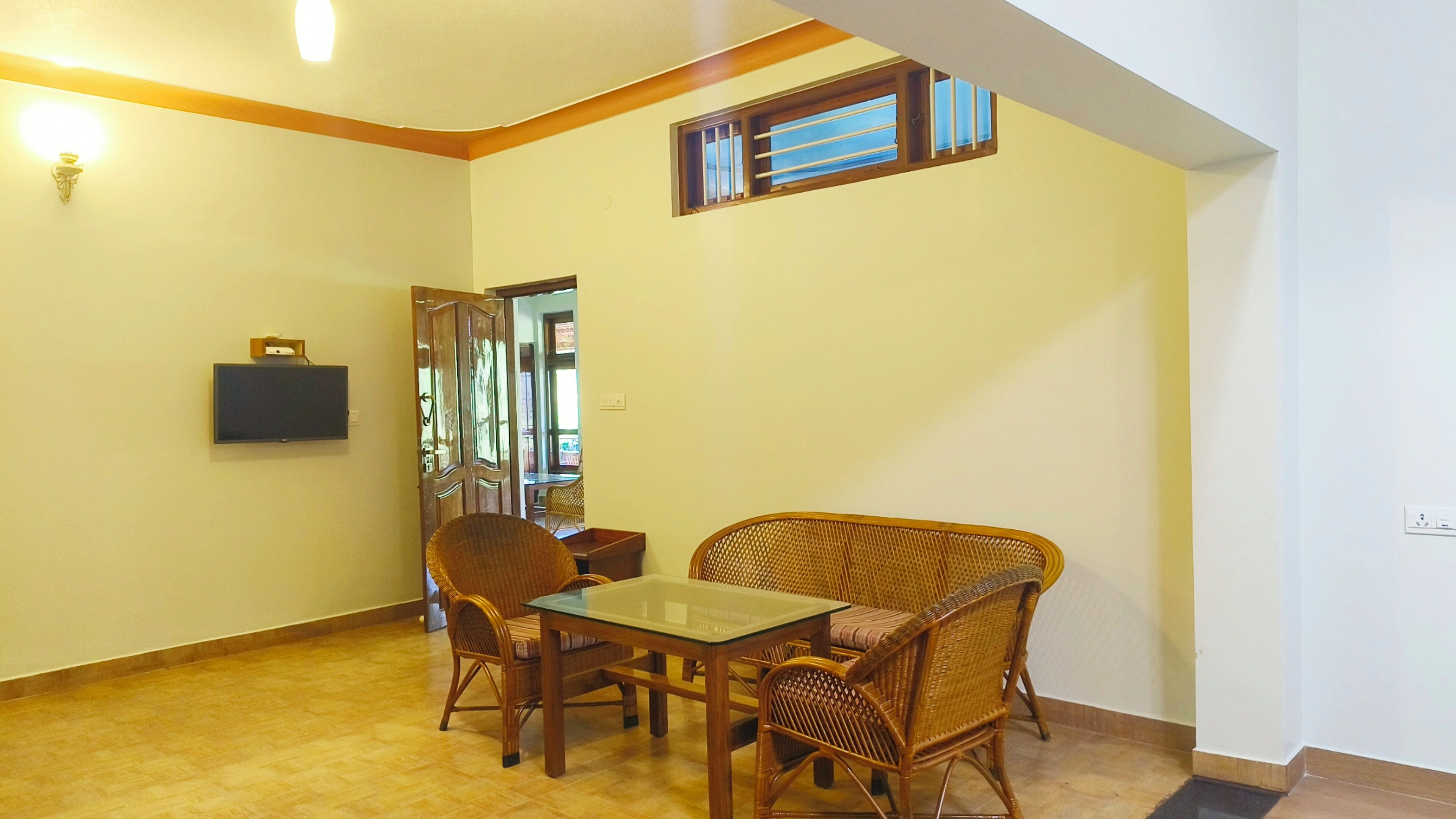 A dining area with a glass-top table and wicker chairs, a flat-screen TV mounted on the wall, and a window with bars.