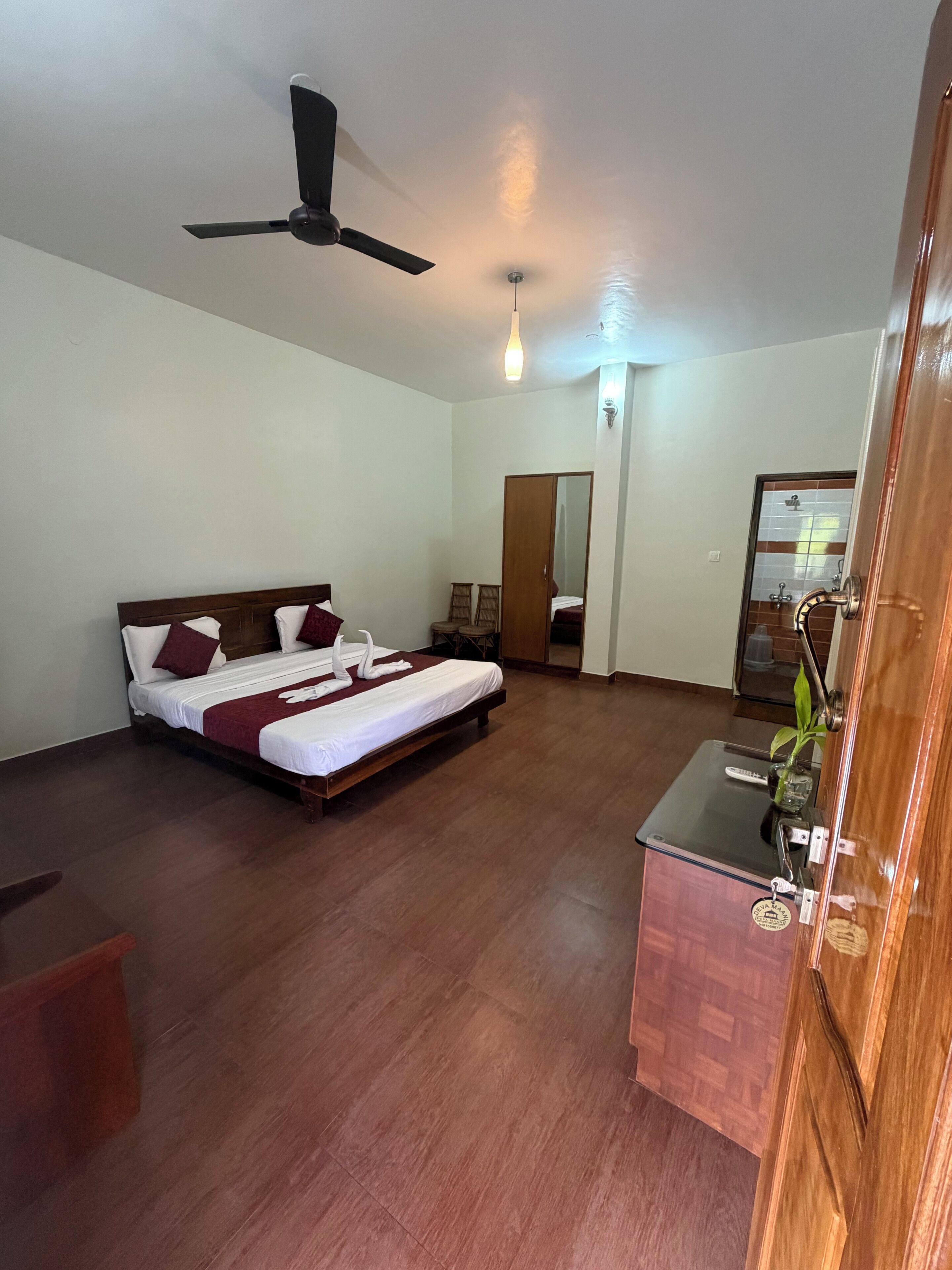A hotel room with a bed, a ceiling fan, a bedside table with a plant, and a door leading to a bathroom.
