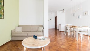 A modern living room with a beige sofa, a round wooden table, and a dining area with white chairs and a wooden table.