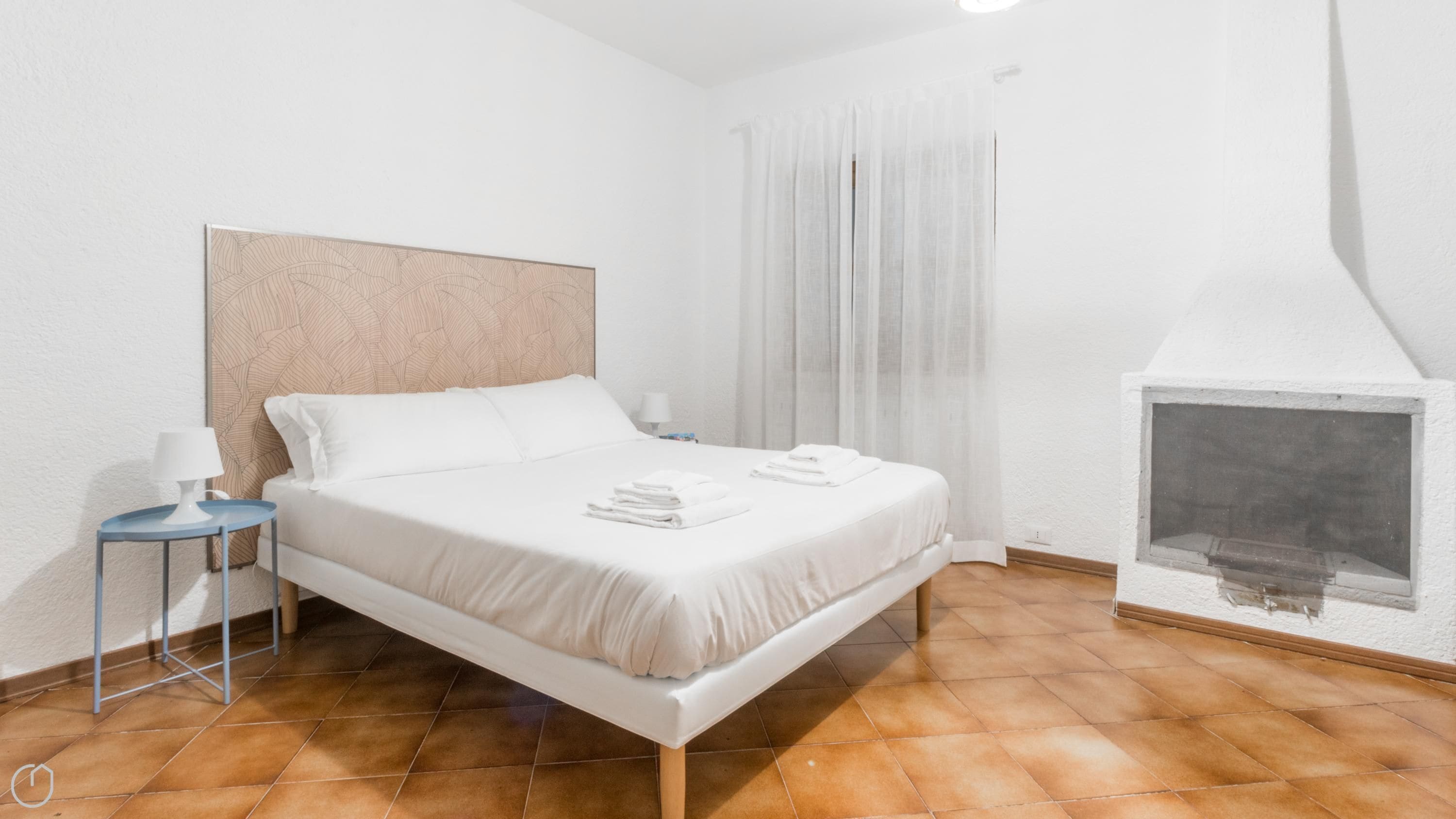 A hotel room with a bed, a nightstand, a lamp, a fireplace, and a window with curtains.