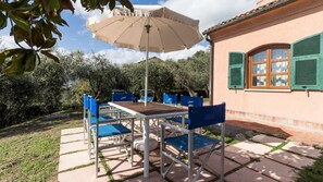 An outdoor patio with a table and chairs, a beige umbrella, and a building with green shutters.