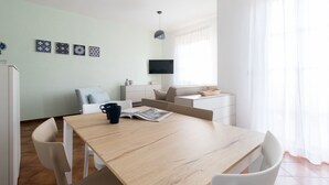 A modern, well-lit room with a wooden dining table, white chairs, a flat-screen TV mounted on the wall, and a window with sheer curtains. - Il Borgo Apartments A3 - Sv-d600-bove3c1a (Finale Ligure)