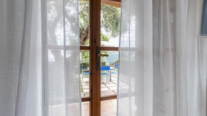 Une vue à travers une fenêtre ornée de rideaux légers, dévoilant un espace extérieur aménagé avec des chaises bleues et une table, ainsi qu’une vue sur un arbre et un bâtiment en arrière-plan.