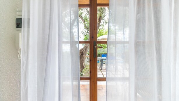 Une pièce avec une porte en bois et des rideaux blancs, un mur blanc, et une vue sur une terrasse extérieure avec un arbre et une table.