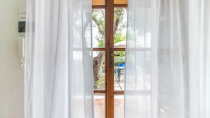 Une pièce avec une porte en bois et des rideaux blancs, un mur blanc, et une vue sur une terrasse extérieure avec un arbre et une table.