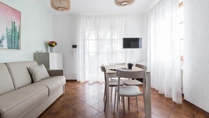Un salon comprenant un canapé, une table à manger avec des chaises, un téléviseur fixé au mur et une fenêtre avec des rideaux.