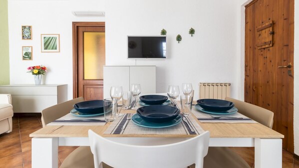 A dining area with a wooden table set for four, surrounded by white chairs. A flat-screen TV is mounted on the wall above the table.