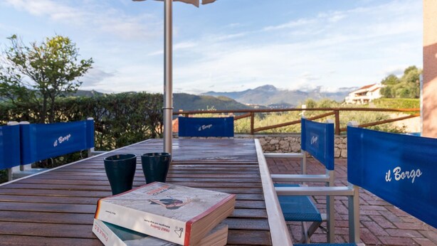 A wooden deck with blue chairs and a table, a book titled "Climbing," and a view of mountains and vineyards. - Il Borgo Apartments C1 - Sv-d600-navi44atc (Finale Ligure)