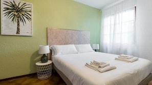 A hotel room with a bed, two bedside lamps, a nightstand, a wall painting, and a window with curtains.