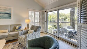 A living room with a sofa, two patterned armchairs, a coffee table, and a lamp. There is a large window with shutters, a painting, and a view of a water body and trees.