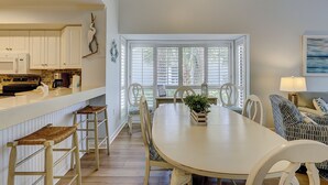 A dining area with a white table, wicker chairs, and a view through large windows.