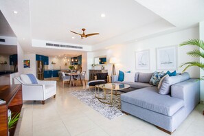 A spacious living area with a grey sofa, a white armchair, and a coffee table. There is a dining area with a table and chairs, and a kitchen in the background.