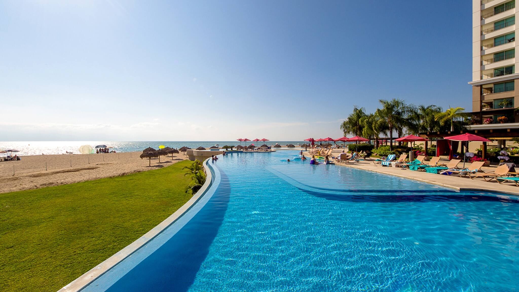 A resort pool with a view of the beach and ocean, surrounded by lounge chairs and umbrellas.