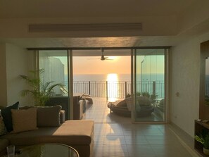 Sala de estar con vista al océano al atardecer, ventilador de techo y puerta de vidrio que da a un área de estar exterior.