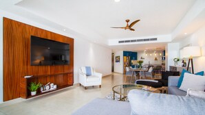 Ein modernes Wohnzimmer mit Flachbildfernseher, Holzpaneelen, Deckenventilator, Essbereich mit Tisch und Stühlen sowie einer Couch mit Kissen.