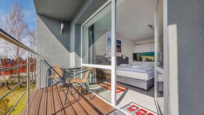 Ein Balkon mit Holzdeck, Korbstühlen und einer Glastür zu einem Schlafzimmer mit Bett, Nachttisch und Gemälde.