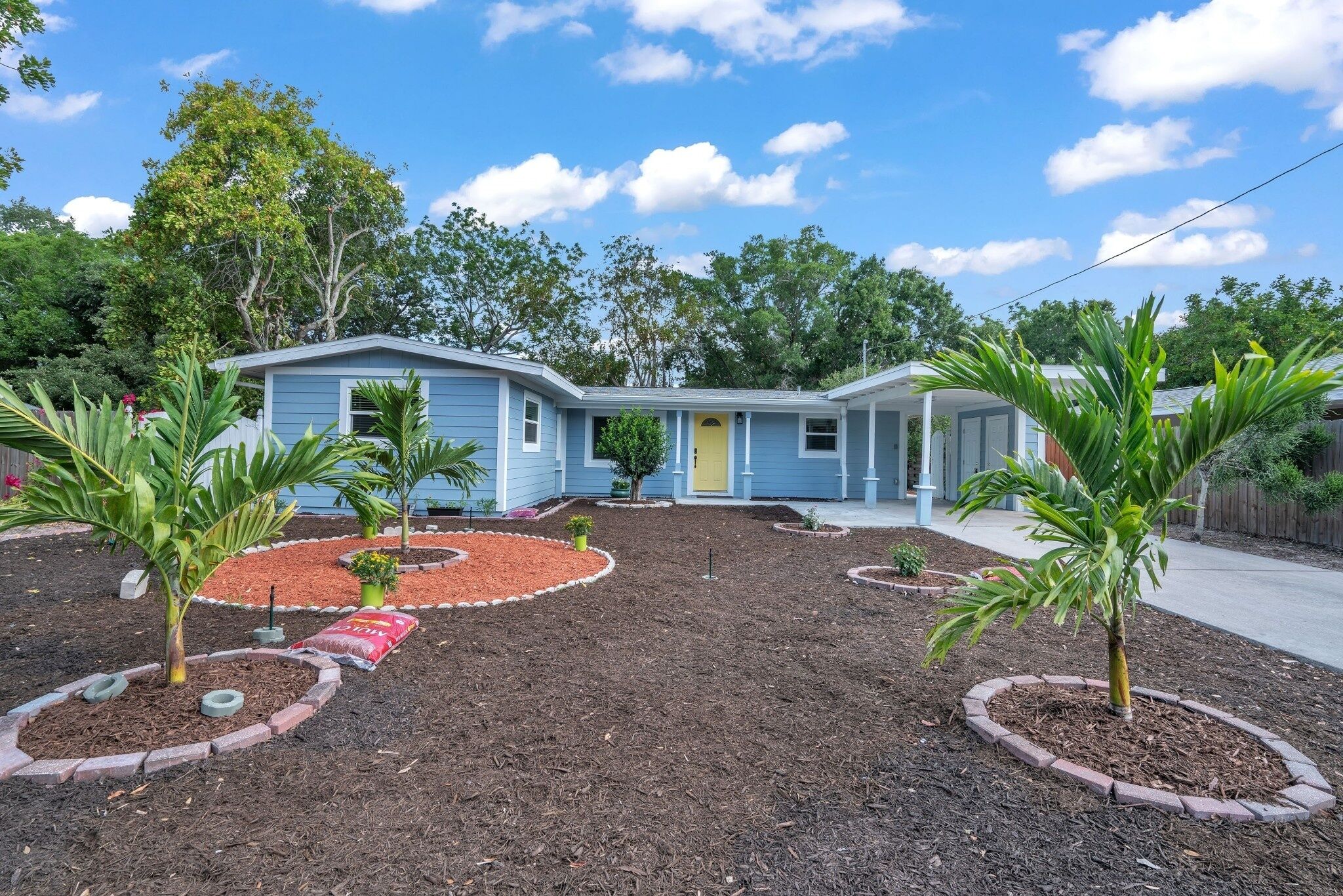 A two-story house with a blue exterior, surrounded by landscaped gardens with palm trees and a circular flower bed.