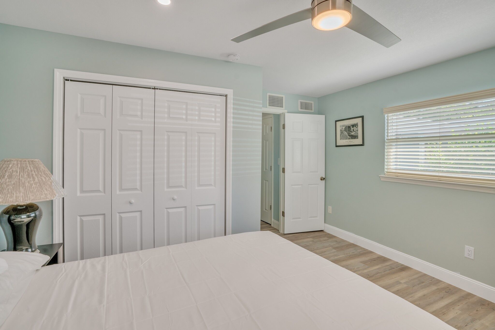 A bedroom with a bed, a ceiling fan, a window with blinds, and a door leading to another room.