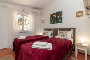 A hotel room with two beds, a window with curtains, a painting on the wall, and a small table with a lamp.
