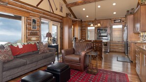 Un salon chaleureux avec un canapé, un fauteuil et un pouf, un coin repas et une cuisine dotée de meubles en bois et d’une grande fenêtre.