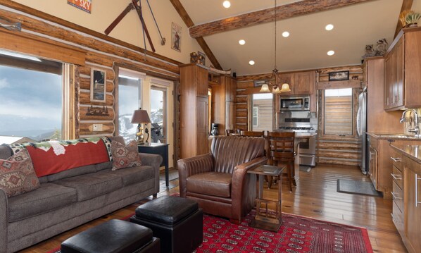 Un salon chaleureux avec un canapé, un fauteuil et un pouf, un coin repas et une cuisine dotée de meubles en bois et d’une grande fenêtre.