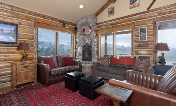 A log cabin interior with a stone fireplace, leather furniture, and large windows offering mountain views.
