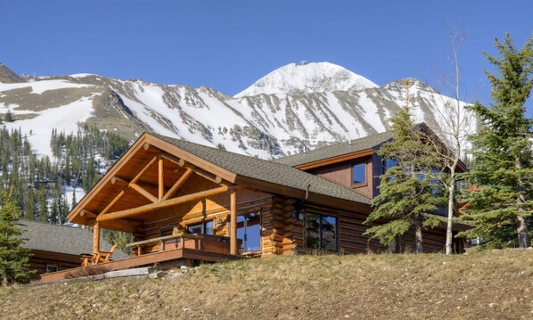 Une cabane en rondins dotée d’une terrasse en bois et de grandes fenêtres, entourée de montagnes et d’arbres couverts de neige.