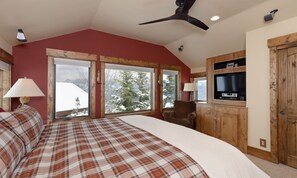 A bedroom with a large bed, two windows with views of snow-covered trees, a ceiling fan, and a TV mounted on a wooden cabinet.
